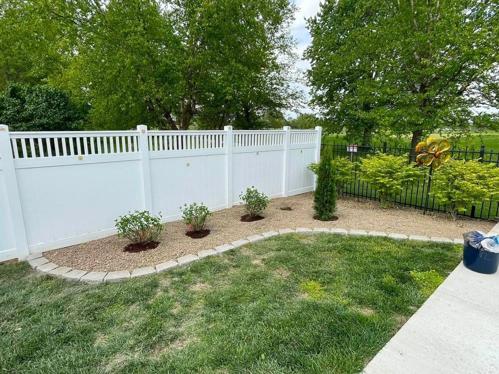 Lush backyard with white vinyl fence, flower beds, and gravel landscaping.
